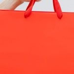 Clearance Sale - Crop unrecognizable female showing red paper shopping bag in hand against white wall